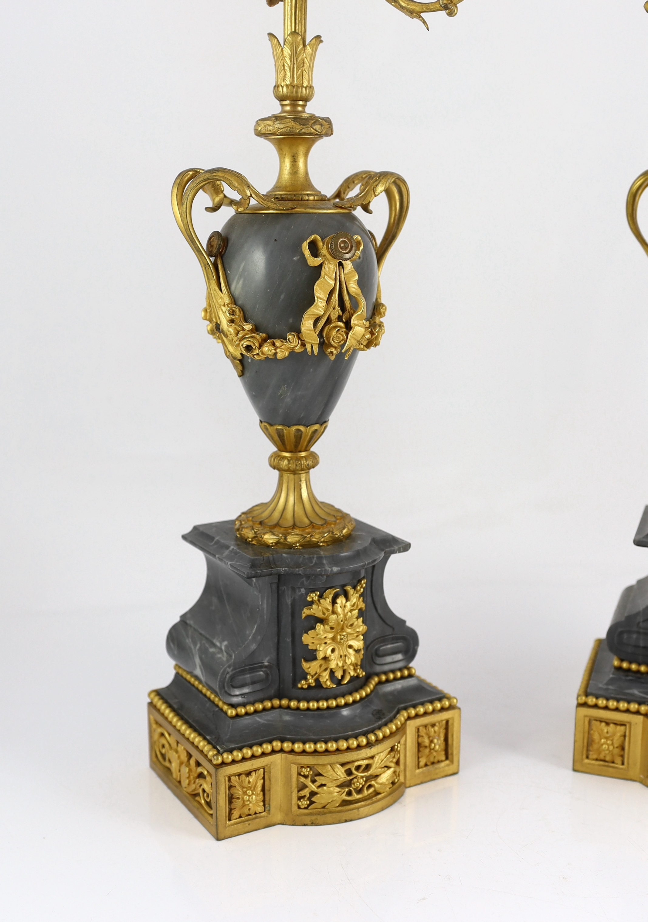 A pair of Louis XVI style ormolu and grey marble five light candelabra, with scrolling branches and urn shaped stems on architectural plinths, 26cm wide, 63cm high